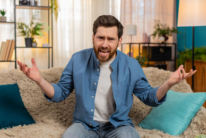 A man sitting on a couch, looking angry and shrugging, representing the frustration caused by a demanding MIL kicking a senior dog. A man sitting on a couch, looking angry and shrugging, representing the frustration caused by a demanding MIL kicking a senior dog.