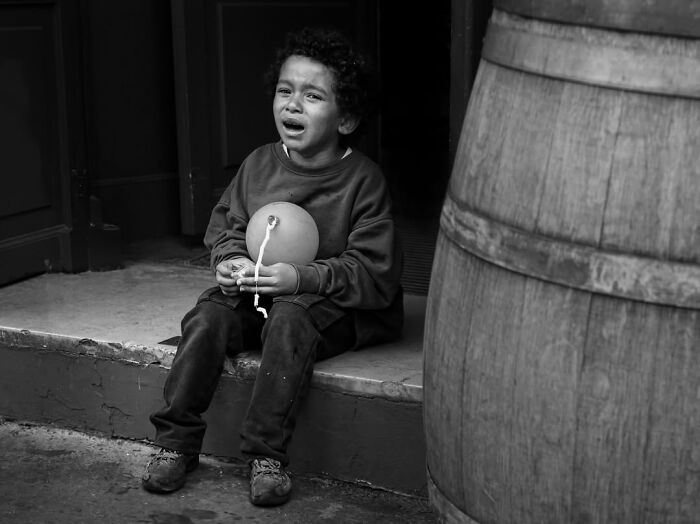 A black and white street photo by Gaspard Claude of a child in Paris crying and holding a balloon while sitting on steps next to a barrel.