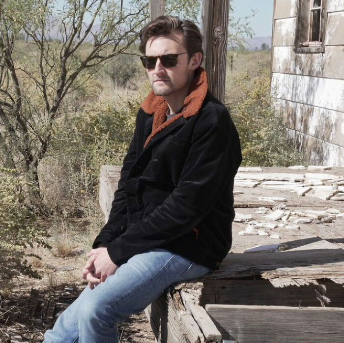 A working class man in sunglasses and a jacket sits on a wooden structure, representing one America.