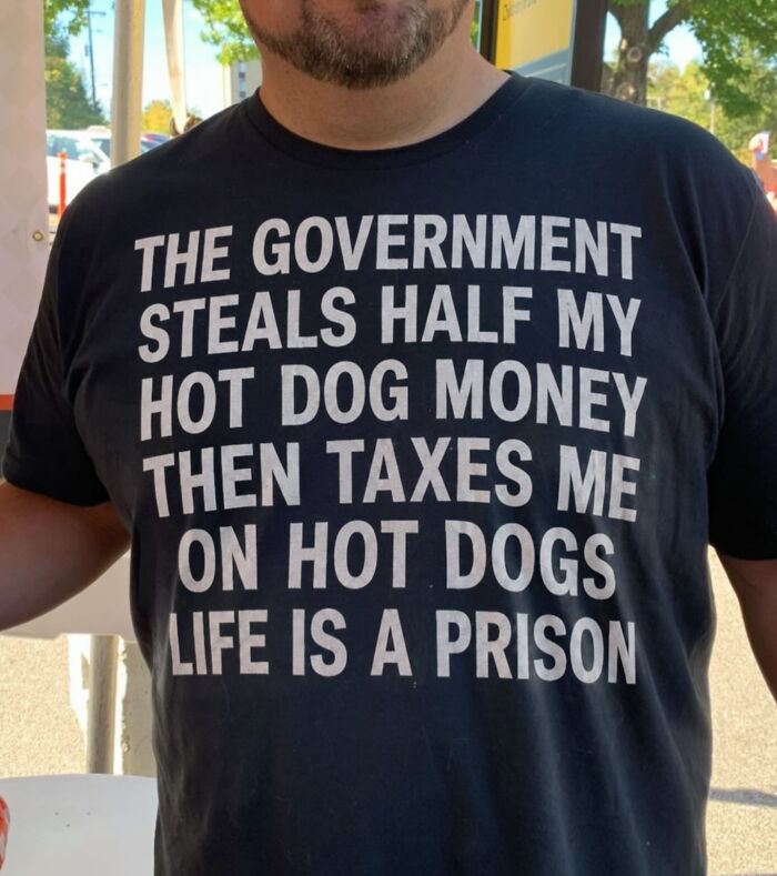 A man wearing a black iconic shirt with white text: THE GOVERNMENT STEALS HALF MY HOT DOG MONEY THEN TAXES ME ON HOT DOGS LIFE IS A PRISON.