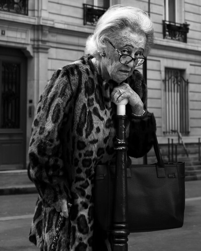 A black and white street photo by Gaspard Claude of an elderly woman in Paris wearing a leopard print coat and glasses, holding a cane.