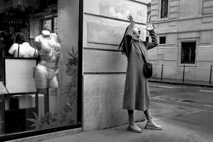 A black and white street photo by Gaspard Claude of a woman in Paris yelling or singing on a street next to a lingerie mannequin.