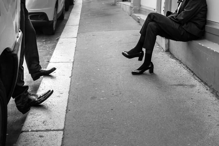 Two pairs of legs on a Paris street, one sitting on a curb and another by a car, a perfectly timed street photo by Gaspard Claude.