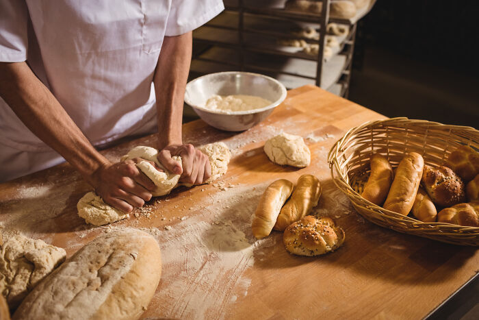 A baker kneading dough on a floured wooden table, surrounded by fresh bread and rolls, showcasing a life-changing skill.