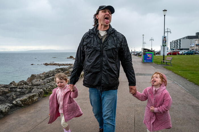 A man holding hands with two children, all sticking out their tongues while walking by the sea, a funny street photography moment.