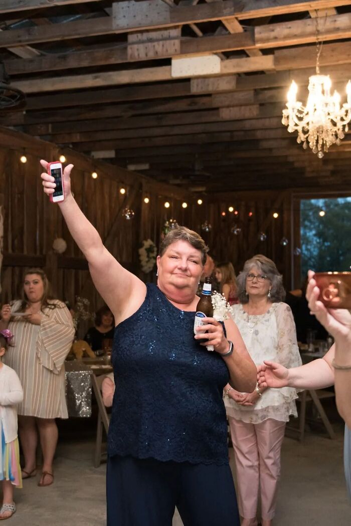 A woman in a navy dress holds a beer and phone aloft on the dance floor, making funny wedding photos.