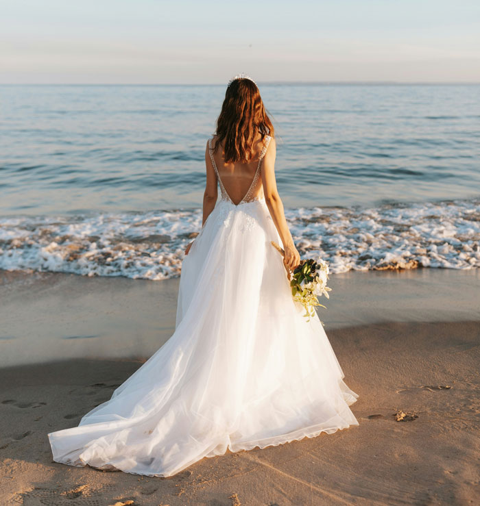 A bride in a white wedding gown, holding a bouquet, stands on a sandy beach looking out at the ocean, pondering if she is wife material.