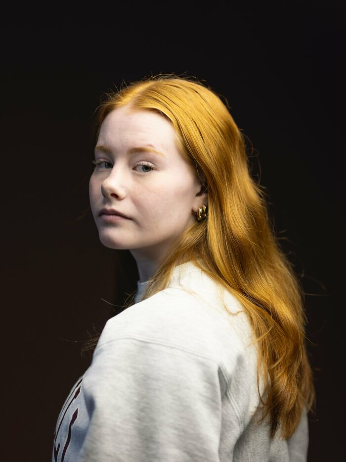 A young woman with long, flowing natural redheads hair and a serious expression, looking over her shoulder.