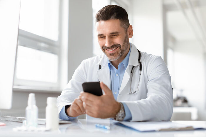 A smiling doctor looking at his phone, showing things doctors refuse to do in their practice.
