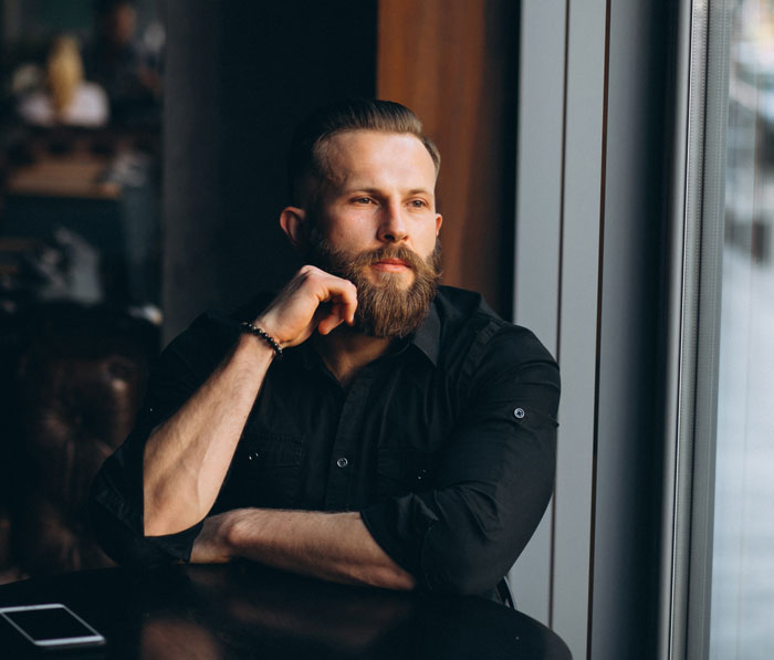 A thoughtful man with a beard looks out a window. He represents a colleague dealing with a workplace bully. A thoughtful man with a beard looks out a window. He represents a colleague dealing with a workplace bully.