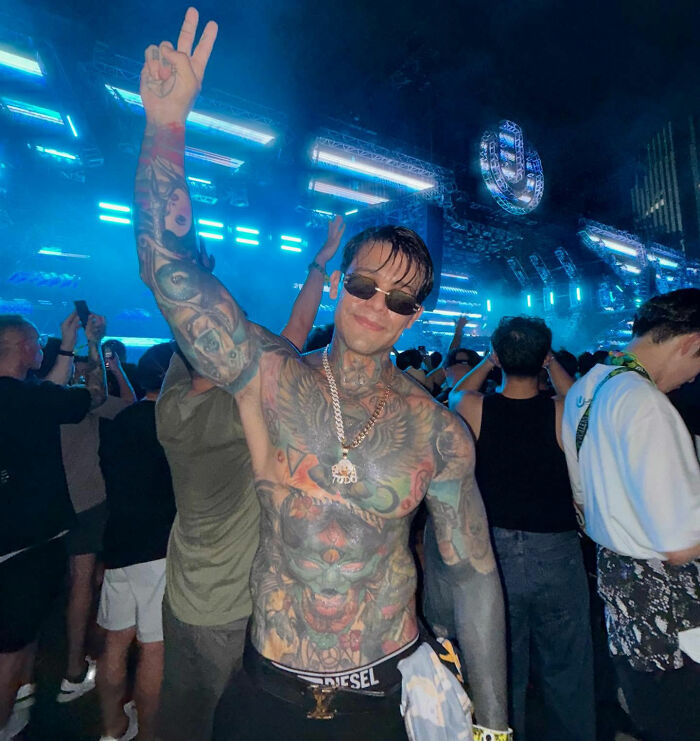Tattooed influencer smiling and giving a peace sign at a crowded event, showcasing his style.
