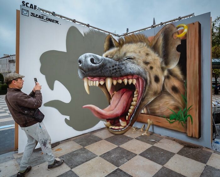 Man photographing a French artist's mind-bending 3D illusion of a hyena bursting from a wall, on a checkered floor.