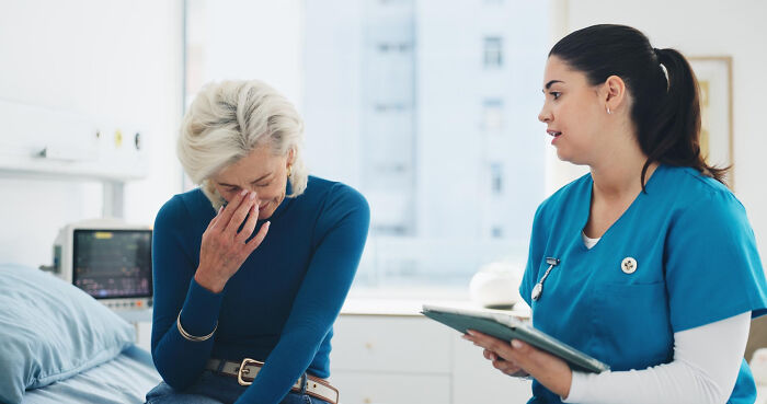 A distressed elderly woman covers her face while a nurse speaks to her, possibly revealing a secret.
