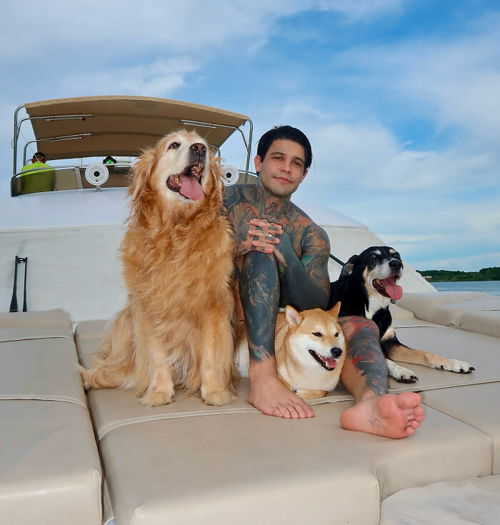 An influencer with many tattoos, sitting on a boat with three dogs, after his mid-flight prank.
