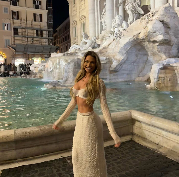 An influencer posing in a white knit outfit in front of the illuminated Trevi Fountain at night, amidst a crowd. An influencer posing in a white knit outfit in front of the illuminated Trevi Fountain at night, amidst a crowd.