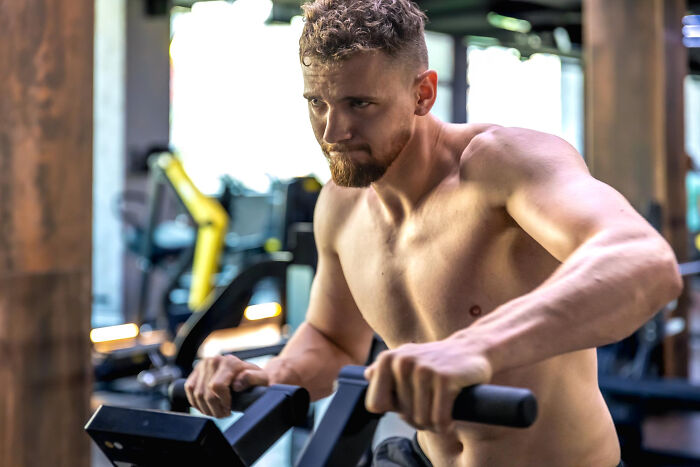 A shirtless man with a beard intensely exercising on a machine in a gym, representing dumb reasons couples divorce.