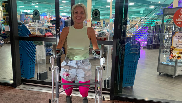 A woman with amputated legs and hands stands with a walker outside a store, showing her reaction after surgery. A woman with amputated legs and hands stands with a walker outside a store, showing her reaction after surgery.
