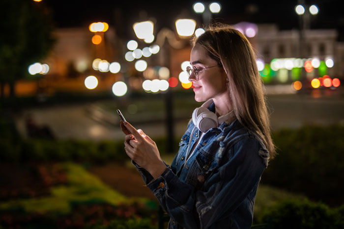 A young woman in glasses and headphones smiles while looking at her phone at night. Women safety tips. A young woman in glasses and headphones smiles while looking at her phone at night. Women safety tips.