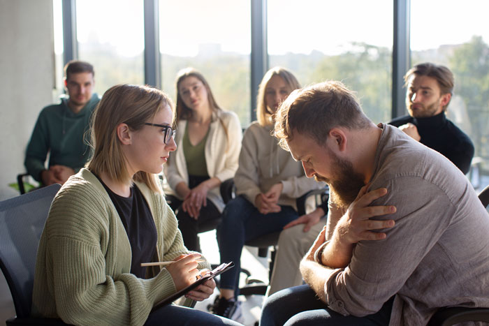 A man with a beard sits slumped, being consoled by a woman in a group therapy setting after losing his job.