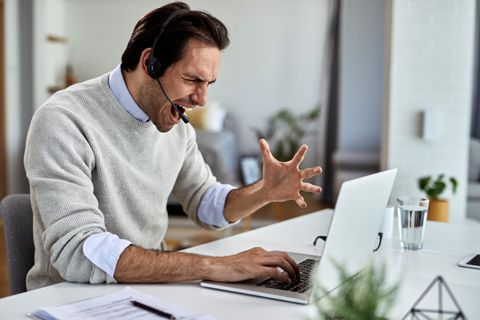 A man wearing a headset, yelling at his laptop, visually representing the frustration caused by a workplace bully stealing credit. A man wearing a headset, yelling at his laptop, visually representing the frustration caused by a workplace bully stealing credit.