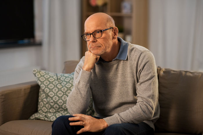 A concerned dad, wearing glasses and a grey sweater, sits on a couch, reflecting on his kids' internships. A concerned dad, wearing glasses and a grey sweater, sits on a couch, reflecting on his kids' internships.