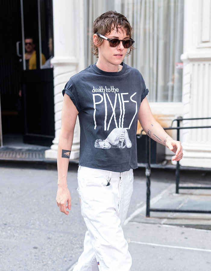 Kristen Stewart on the street, wearing sunglasses, a Pixies T-shirt, and white pants, reflecting her unsettling appearance.