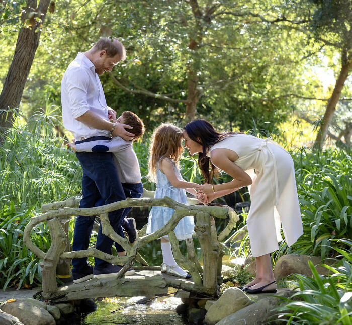 Prince Harry, Meghan Markle, and their children on a rustic wooden bridge in a lush green garden. Prince Harry, Meghan Markle, and their children on a rustic wooden bridge in a lush green garden.