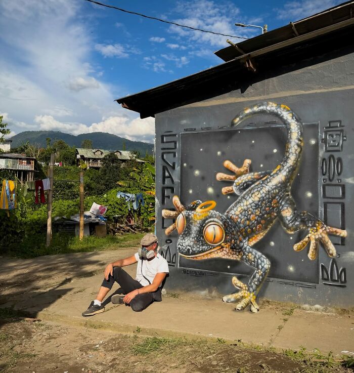 French artist next to a mind-bending 3D illusion of a spotted gecko on an ordinary wall in a rural setting.