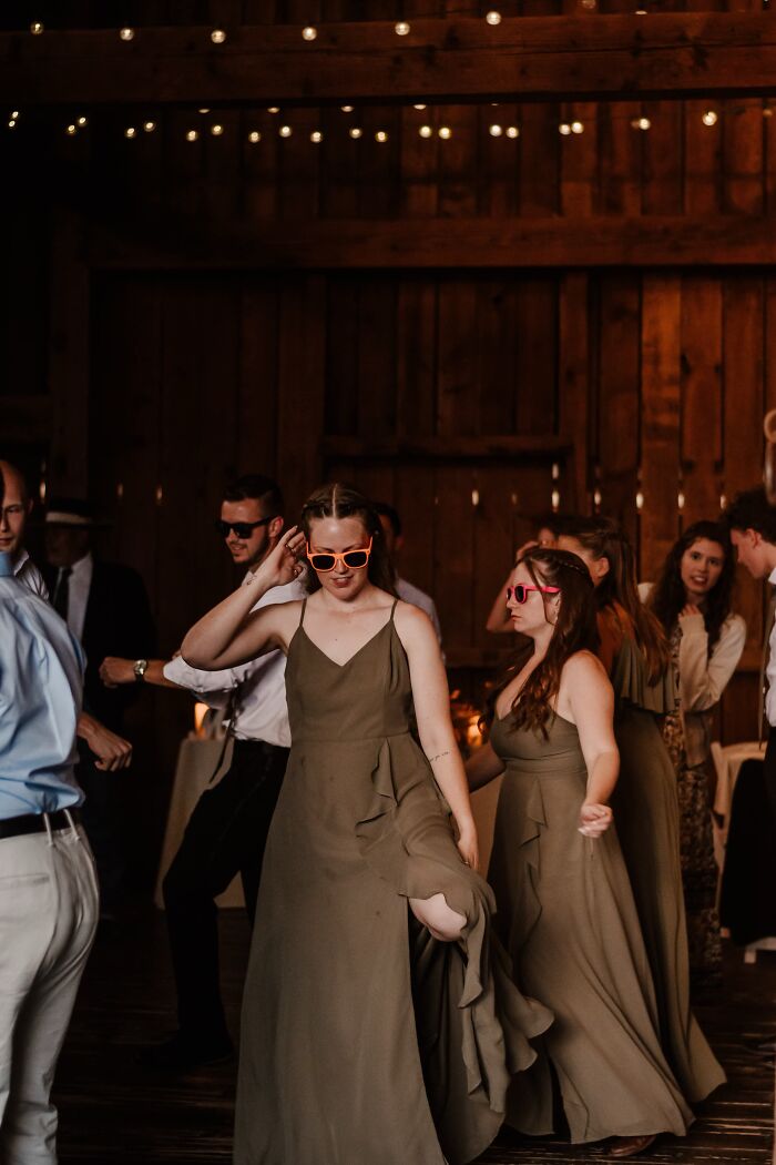 A funny wedding photo of bridesmaids wearing pink heart-shaped sunglasses and dancing at a rustic wedding reception.