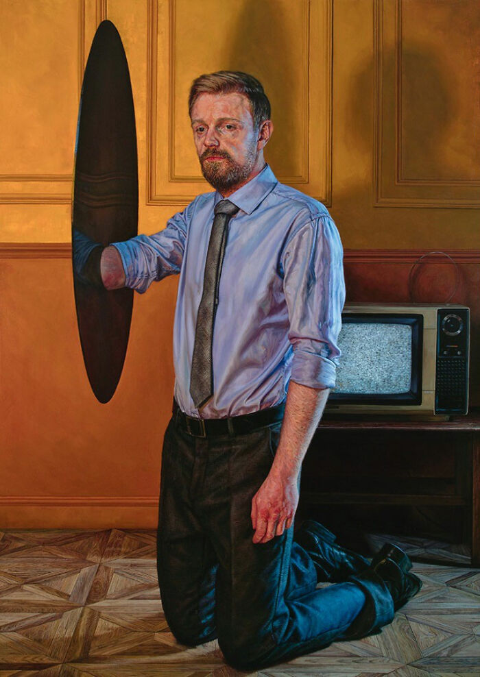 Hyperrealist paintings by Ian Cumberland: a man in a blue shirt and tie kneels, holding a dark oval.