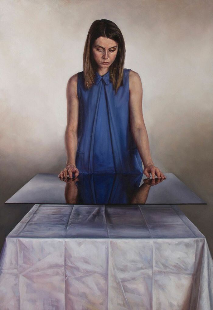 A hyperrealist painting by Ian Cumberland showing a woman in blue looking at her reflection on a table.