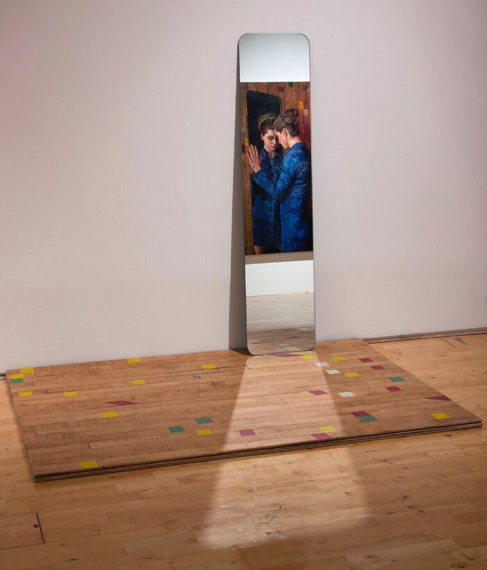 A hyperrealist painting by Ian Cumberland showing a woman reflected in a tall mirror with surreal details.