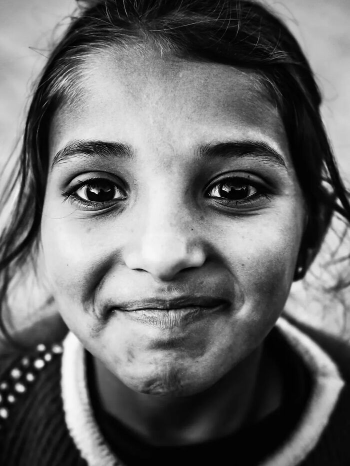 A perfectly timed street photo by Mehmet Bayram, a close-up, black and white portrait of a young girl with a gentle smile.