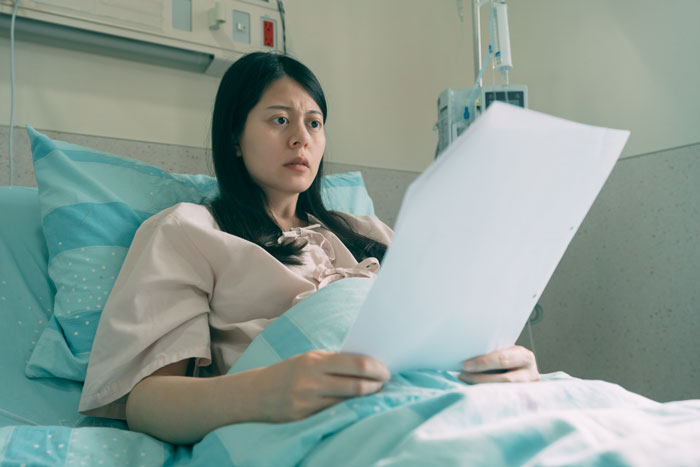 A worried woman in a hospital bed holding papers, facing a costly price after donating a kidney. A worried woman in a hospital bed holding papers, facing a costly price after donating a kidney.