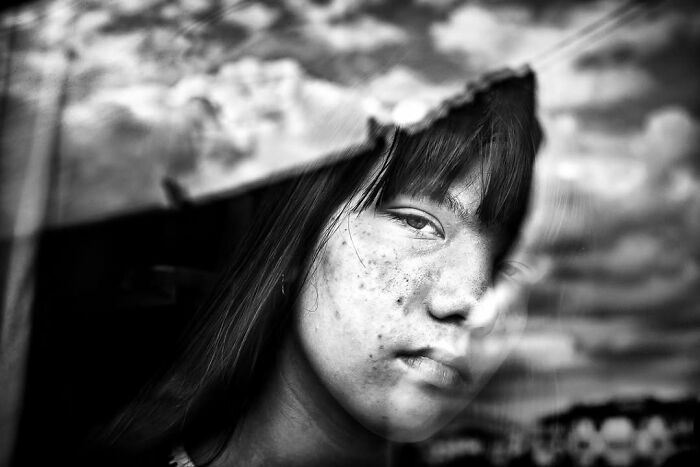 A black and white street photo by Murat Harmanlikli capturing the curious face of a child with freckles and bangs.