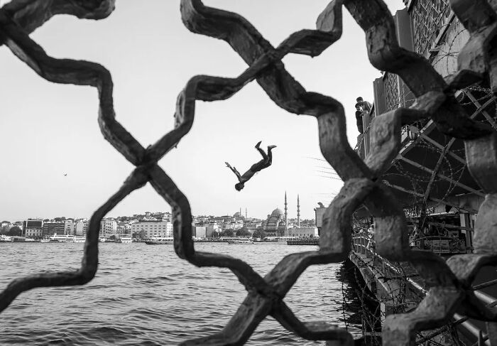 Candid moments from the streets, a person diving into the sea, framed by an ornate metal fence, with a city skyline in the background in black and white.