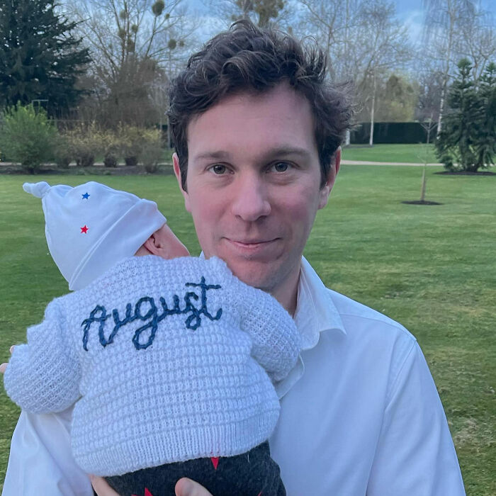 A man holding a baby wearing a knitted sweater that says August. This image relates to Meghan Markle's karma. A man holding a baby wearing a knitted sweater that says August. This image relates to Meghan Markle's karma.