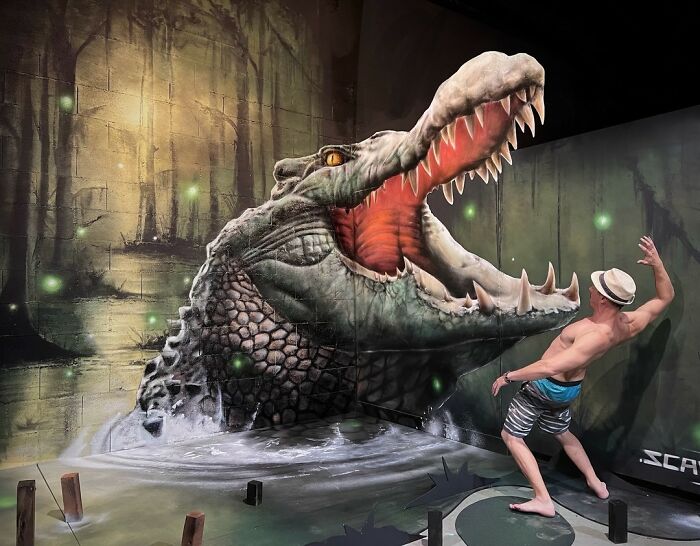 French artist in a hat posing with a mind-bending 3D illusion of an alligator emerging from a swamp.