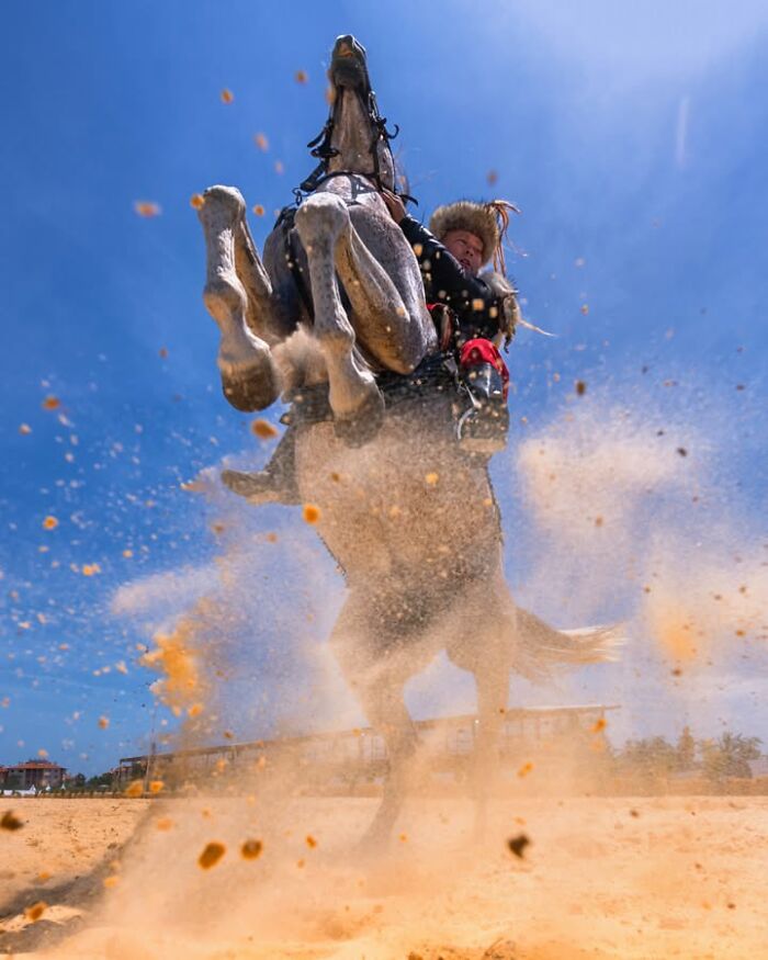 Murat Bakmaz captures the beauty of everyday life: A rider on a horse rearing up, kicking up dust under a clear blue sky.