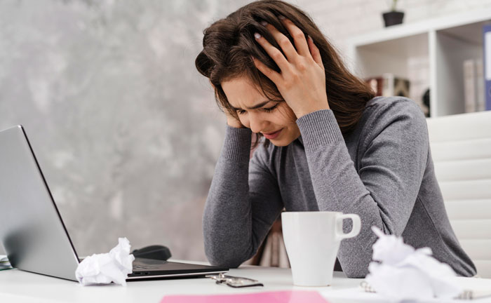A distressed woman holding her head at a desk with a laptop and coffee, reflecting the dad's dilemma with kids internships. A distressed woman holding her head at a desk with a laptop and coffee, reflecting the dad's dilemma with kids internships.