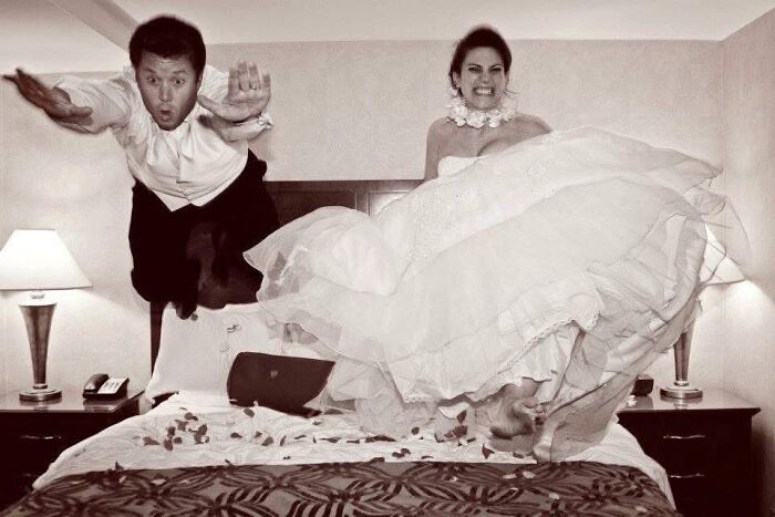 Iconic funny wedding photo of a newlywed couple mid-air jumping on a bed in their wedding attire.