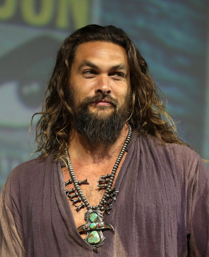 Jason Momoa with long hair and beard, wearing a rustic shirt and a turquoise necklace. Career highlights for Jason Momoa.