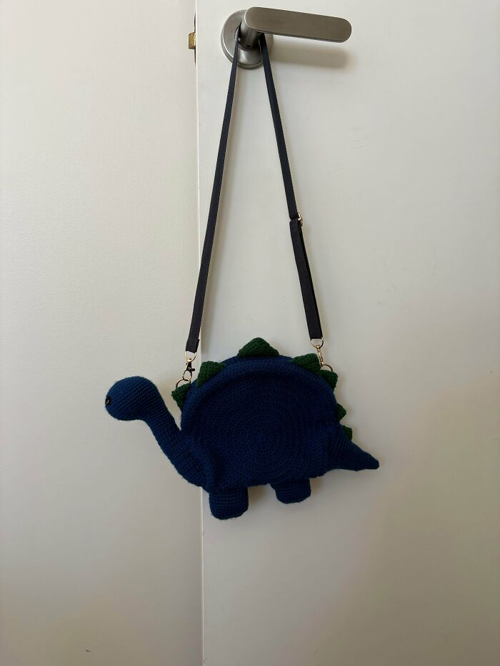 Crochet dinosaur shaped bag hanging from a door handle with adjustable strap