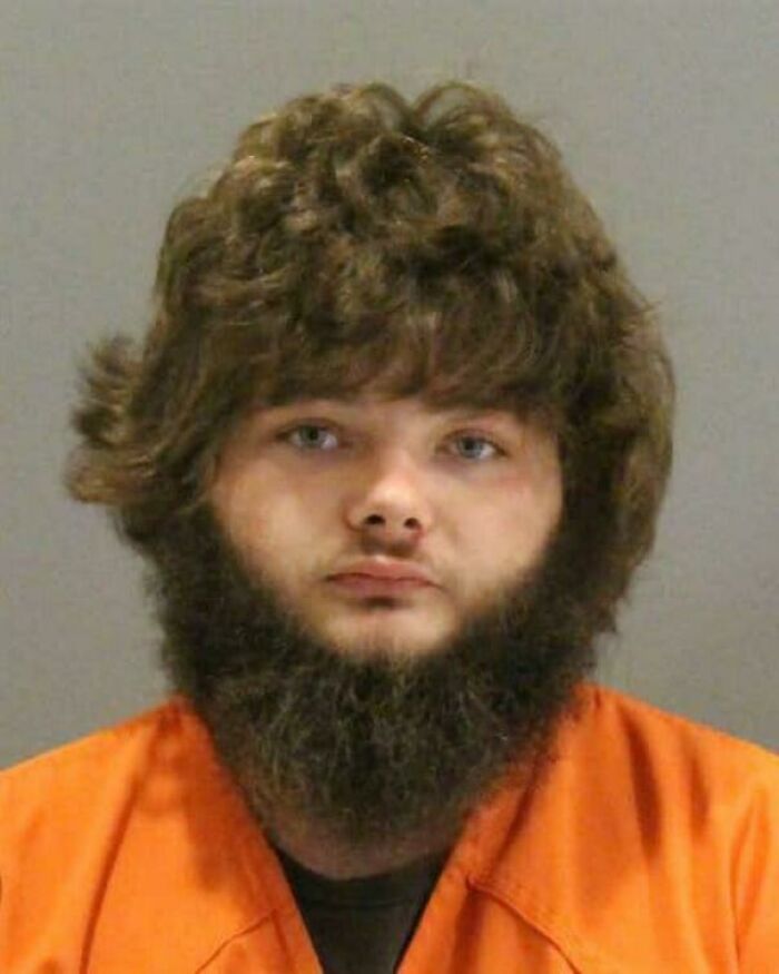 A mugshot of a man with bushy brown hairstyles, a full beard, and light eyes, wearing an orange jumpsuit.