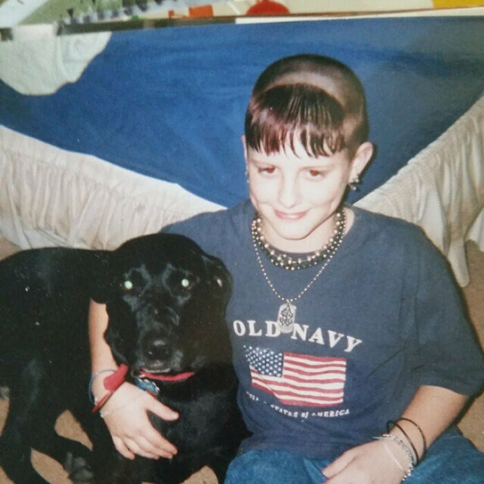 A young person with a shaved head and short bangs, a bold hairstyle, hugging a black dog.