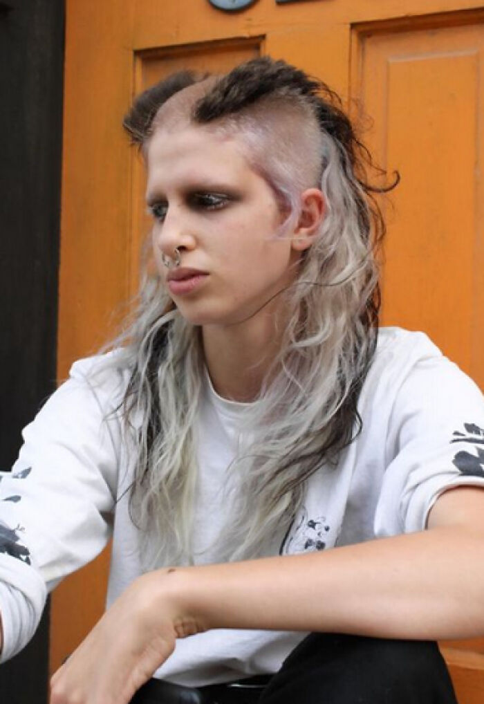 A person with a bold, partially shaved hairstyle and long, streaky hair, featuring a nose ring, against an orange door.