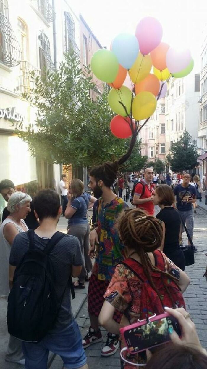 A man with dreadlocks holding a cluster of colorful balloons that appear to be growing from his head, in a street scene.