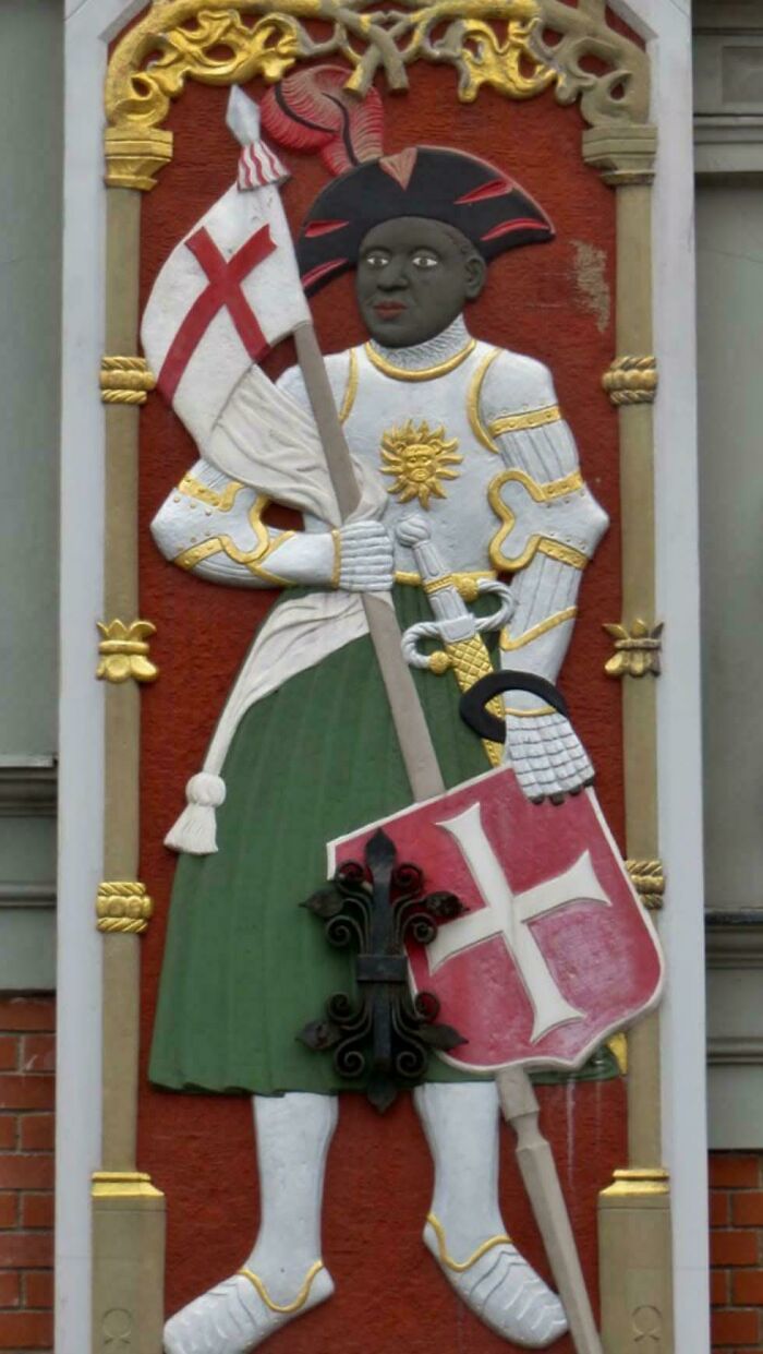 A carved relief of a knight from the Middle Ages, holding a flag and a shield with a red cross.