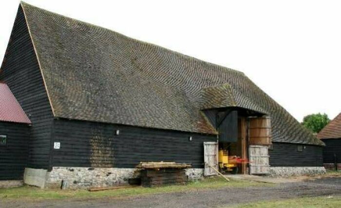 A large, dark wooden barn with a steeply pitched roof, typical architecture from the Middle Ages.