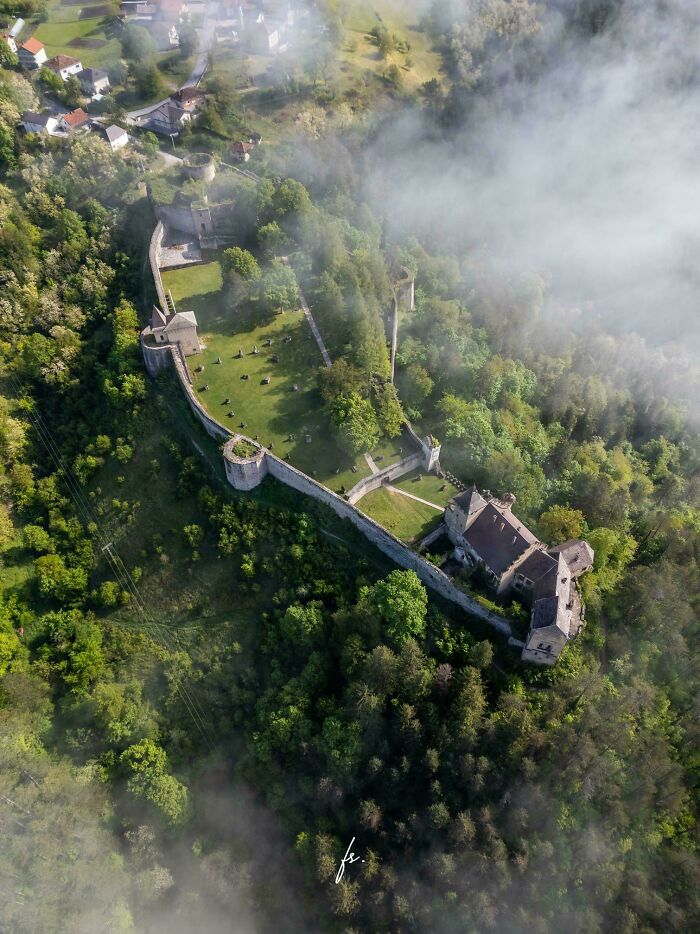 An aerial view of a medieval castle ruin nestled in lush green trees, partially obscured by mist, a fascinating view of the Middle Ages.